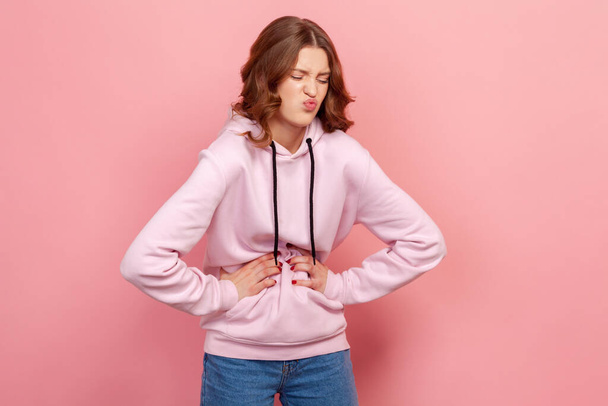 Portrait of upset teenager female in hoodie grimacing and holding belly, suffering abdominal pain and spasm, gastritis. Indoor studio shot isolated on pink background - Foto, immagini