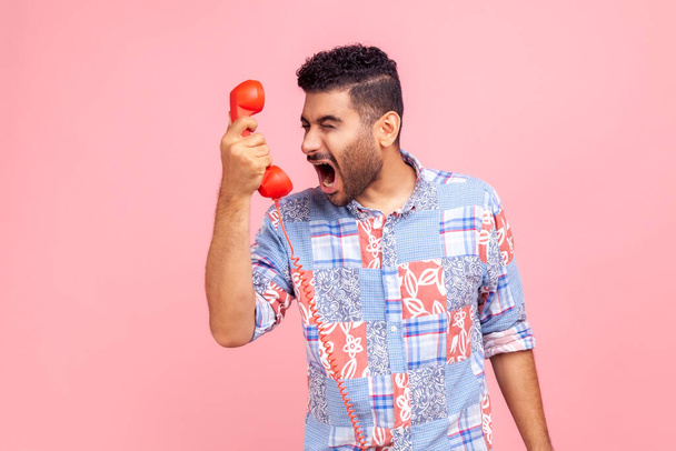 Portrait of bearded angry man screaming into phone handset, annoyed by telephone conversation, aggressive customer calling contact center. Indoor studio shot isolated on pink background. - Foto, Imagen