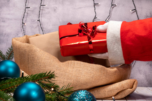 Père Noël prépare le sac de jute avec des cadeaux de Noël. Main avec gant blanc tenant une boîte cadeau avec papier et ruban rouge. - Photo, image