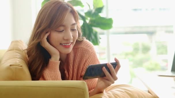 hermosa mujer asiática sonriendo feliz en casa en la sala de estar acostado en el teléfono, haciendo videollamadas en línea, charlando con amigos. Concepto moderno de tecnología de la comunicación, distancia social. - Metraje, vídeo