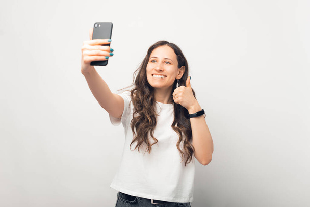Όμορφη γυναίκα παίρνει μια selfie με το τηλέφωνό της, ενώ εμφανίζονται αντίχειρα πάνω από λευκό φόντο. - Φωτογραφία, εικόνα