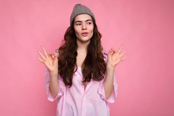 Beautiful pretty nice cute calm concentrated young curly brunette woman wearing pink shirt and grey hat isolated on pink background with copy space. - Photo, Image
