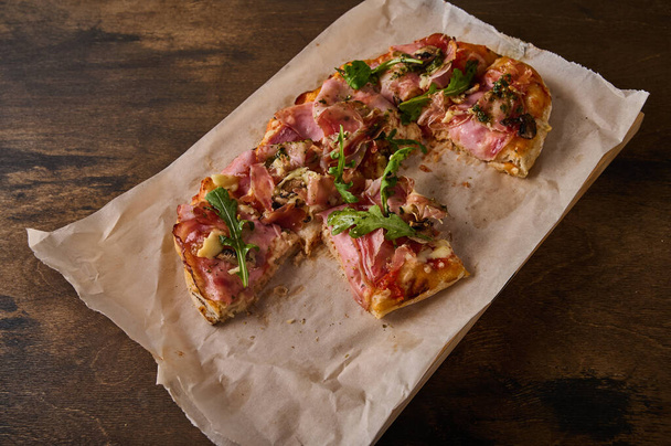 Cut into pieces rectangular Roman pizza with prosciutto ham, tomatoes, mozzarella and arugula on on paper on wooden background - Zdjęcie, obraz