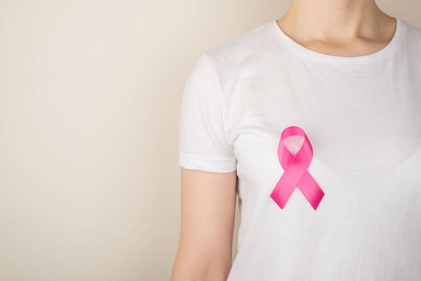 Фото молодой женщины в белой футболке с розовой лентой, символом информированности о раке груди на изолированном сером фоне с копирайтом - Фото, изображение