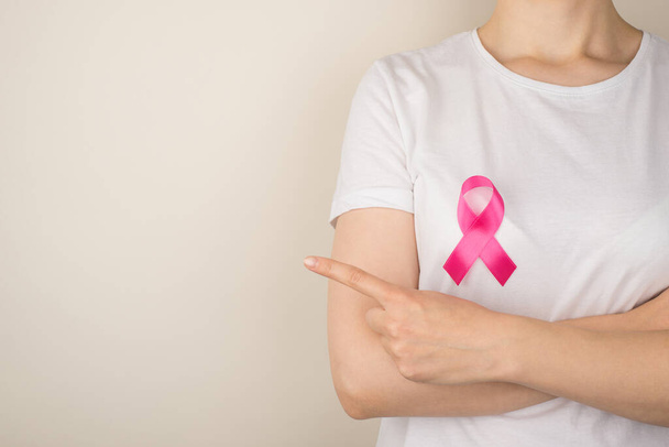 Photo of young woman in white t-shirt with pink ribbon symbol of breast cancer awareness pointing to something with forefinger on isolated grey background with blank space - Photo, Image