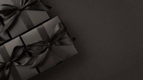 Top view photo of stylish gift boxes in black packaging with tag and black ribbon bow on isolated black background with blank space - Photo, Image