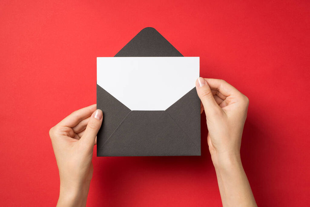 First person top view photo of hands holding open black envelope with white card on isolated red background with blank space - Foto, Imagen