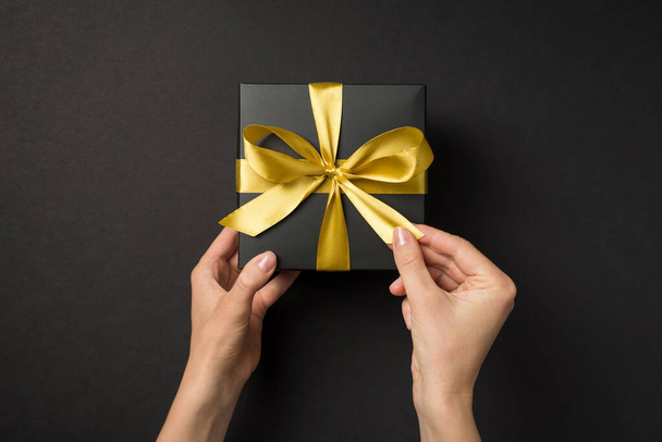 First person top view photo of hands opening black giftbox with golden satin ribbon bow on isolated black background - Foto, immagini