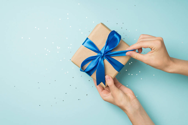 First person top view photo of hands unpacking craft paper gift box with blue satin ribbon bow over shiny sequins on isolated pastel blue background with blank space - Φωτογραφία, εικόνα