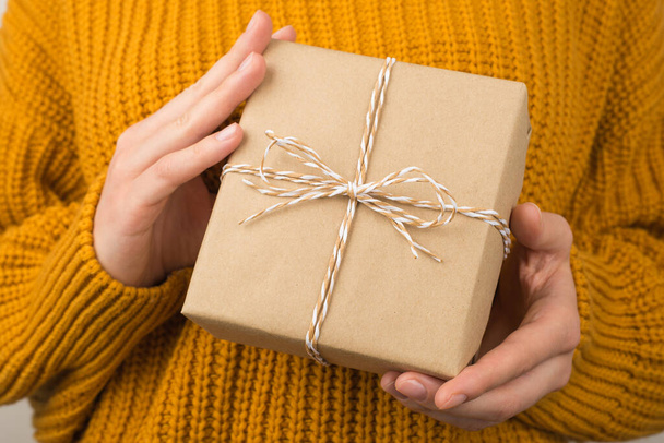 Photo of young woman in yellow cozy sweater demonstrating stylish craft paper giftbox with twine bow - Zdjęcie, obraz