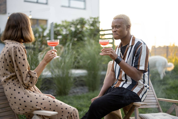 Monirotuinen pari juo ja paahtaa cocktaileja kotijuhlissa puutarhassa. Suhteen käsite. Kotimainen elämäntapa. Musta mies ja eurooppalainen tyttö nauttivat aikaa yhdessä - Valokuva, kuva