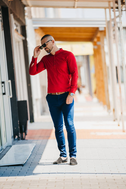 A cool handsome Caucasian man wearing an elegant red shirt and sunglasses posing outdoor - Foto, afbeelding