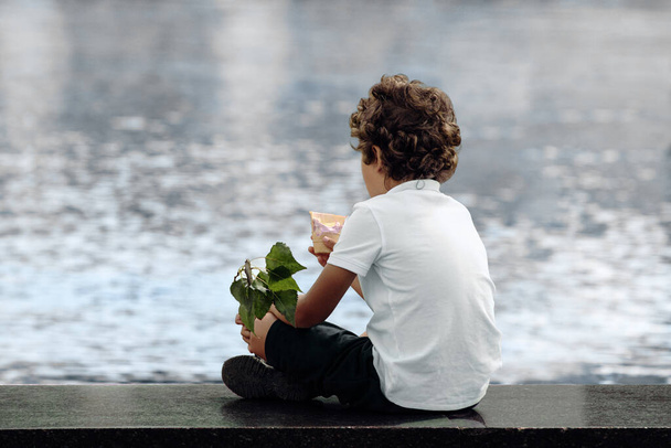 Unrecognizable curly boy holding ice cream cone and green branch in his hands, sitting in city park contemplating water in fountain and dreaming. Rear view - Photo, Image