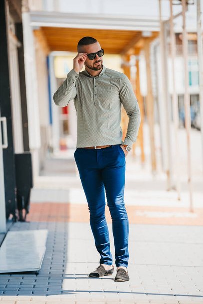 A cool handsome Caucasian man wearing an elegant gray shirt and sunglasses posing outdoor - Photo, Image