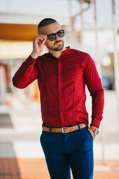 A cool handsome Caucasian man wearing an elegant red shirt and sunglasses posing outdoor - Foto, imagen