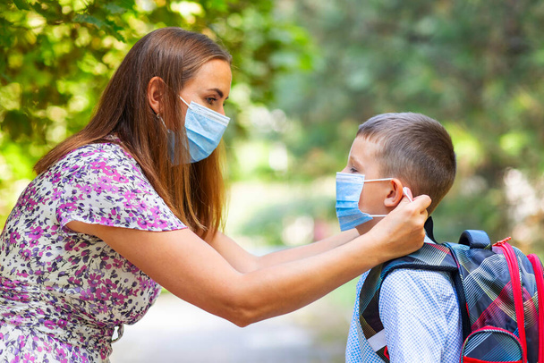 Tıbbi maskeli küçük bir çocuk ve anne. Anne oğullarının yüzüne koruyucu bir maske takıyor. Öğrenci çocuk okula gitmeye hazır. Dışarıda sırt çantası olan sevimli bir çocuk. Okul konseptine dönelim.. - Fotoğraf, Görsel