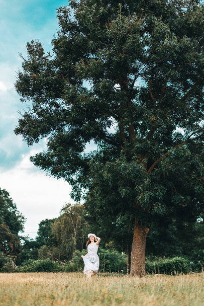 Счастливая молодая женщина в платье и соломенной шляпе бежит через поле. На заднем плане большое дерево и бурное небо. По вертикали. Копирование пространства. - Фото, изображение