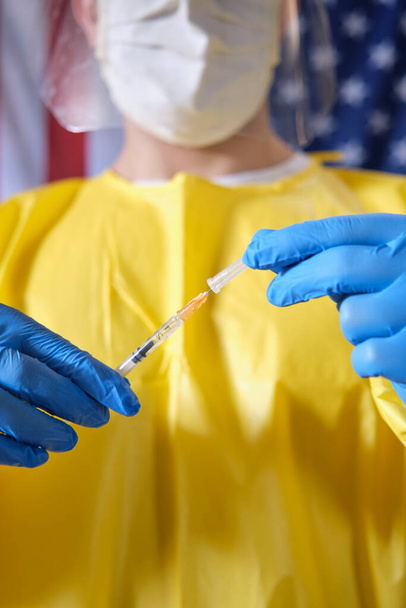 Primer plano El joven trabajador de la salud observa la aguja de una jeringa para inyección con la bandera de los Estados Unidos en el fondo - Foto, imagen