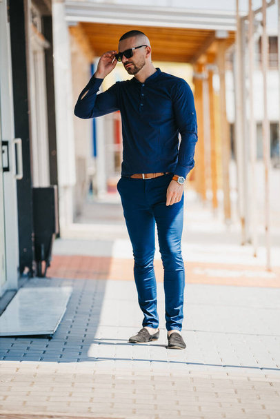 A cool handsome Caucasian man wearing an elegant blue shirt and sunglasses posing outdoor - Zdjęcie, obraz
