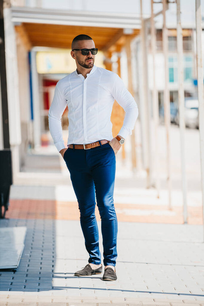 A cool handsome Caucasian man wearing an elegant white shirt and sunglasses posing outdoor - Fotografie, Obrázek