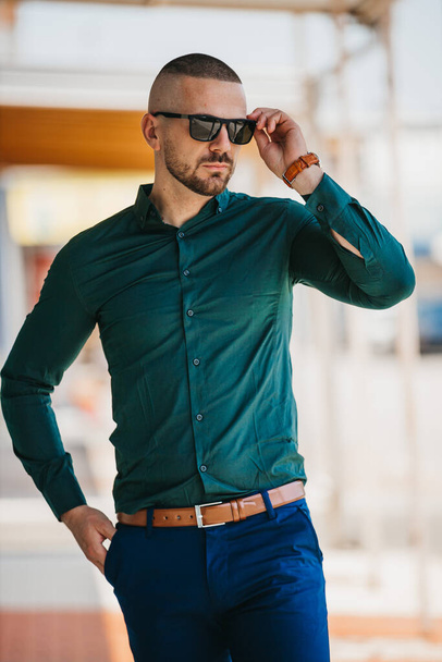 A cool handsome Caucasian man wearing an elegant green shirt and sunglasses posing outdoor - Photo, Image