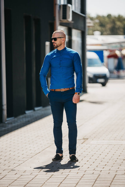 A cool handsome Caucasian man wearing an elegant blue shirt and sunglasses posing outdoor - Foto, Imagem