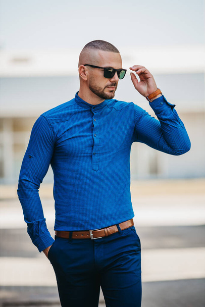 A cool handsome Caucasian man wearing an elegant blue shirt and sunglasses posing outdoor - Фото, изображение