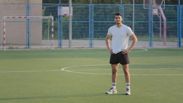 Привабливий спортсмен, індійський хлопець в літній спортивній манері відпочиває між тренуваннями, дихаючи свіжим повітрям. Спортсмен Середнього Сходу робить перерву, радісно посміхаючись, стоячи на футбольному полі стадіону. - Кадри, відео