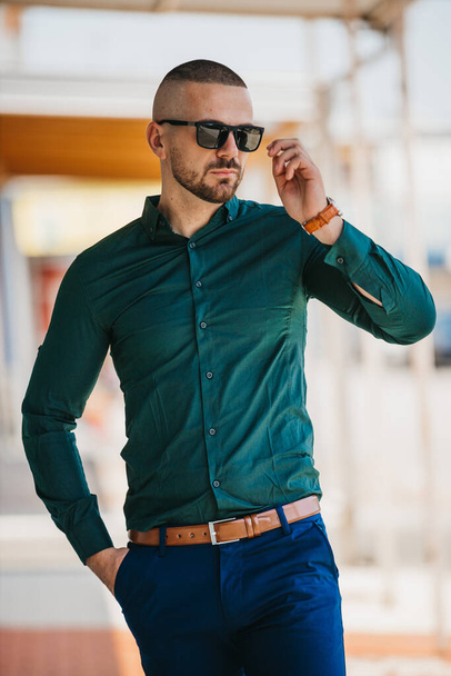 A cool handsome Caucasian man wearing an elegant green shirt and sunglasses posing outdoor - Zdjęcie, obraz