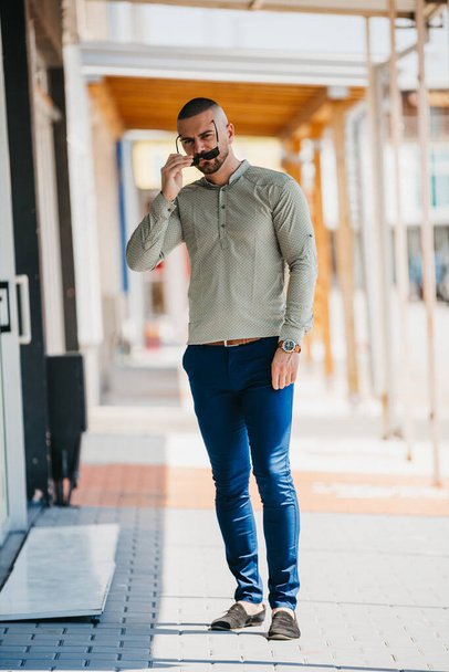 A cool handsome Caucasian man wearing an elegant gray shirt and sunglasses posing outdoor - Fotografie, Obrázek