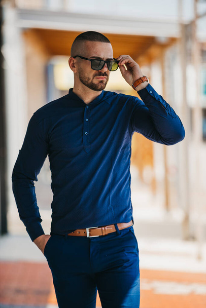 A cool handsome Caucasian man wearing an elegant blue shirt and sunglasses posing outdoor - Zdjęcie, obraz