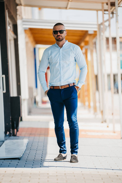 A cool handsome Caucasian man wearing an elegant light blue shirt and sunglasses posing outdoor - Fotografie, Obrázek