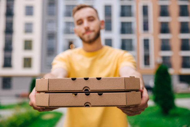 Shooting a portrait of a handsome delivery man standing in the street holding cardboard boxes while handing over the camera. Male courier handing pizza to customer. - Photo, Image