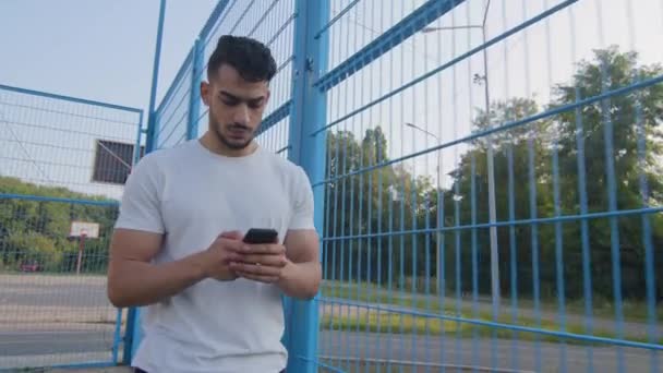 携帯電話を保持してスタジアムフェンスに沿って移動し、スマートフォン上のオンラインアプリを使用して、夏のスポーツウェアの歩行中の若い中東アラブ選手、テキストメッセージを入力チャット、タッチスクリーン - 映像、動画