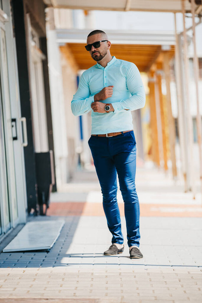 A cool handsome Caucasian man wearing an elegant light blue shirt and sunglasses posing outdoor - Fotó, kép