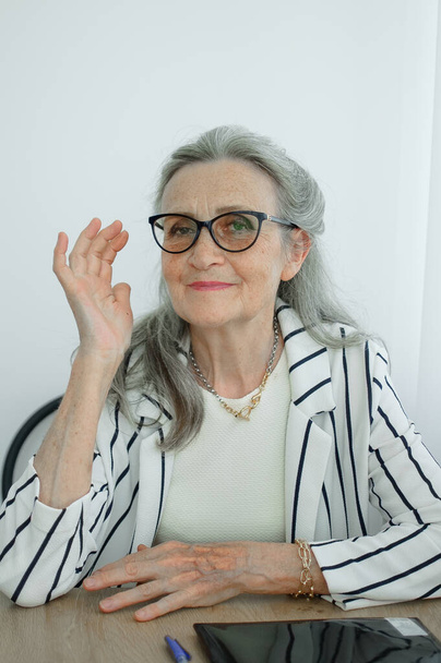 Senior grijs harige zakenvrouw in gestreepte jas met bril werkt in haar kantoor aan het bureau. Gelukkige pensionering, werkgelegenheid en arbeid - Foto, afbeelding