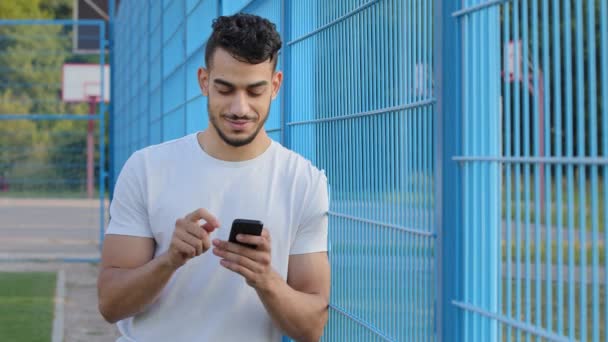 Středovýchodní indián drží telefon v rukou, píše zprávy nebo používá mobilní aplikace, kontroluje aplikace sociálních médií na smartphonu. Mladý arabský sportovec v letním sportovním oblečení hraje na mobilu - Záběry, video