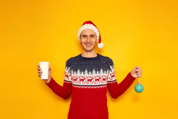 smiling man in Christmas sweater with reindeer, Santa Claus hat holds Christmas tree toy and paper cup coffee, tea take away. isolated yellow background with space for text. concept - holiday, drinks - Photo, Image