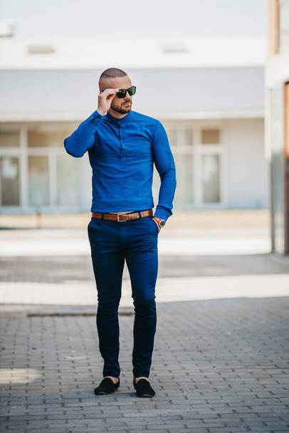Un hombre caucásico guapo fresco usando una camisa azul elegante y gafas de sol posando al aire libre - Foto, imagen