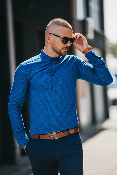 A cool handsome Caucasian man wearing an elegant blue shirt and sunglasses posing outdoor - 写真・画像