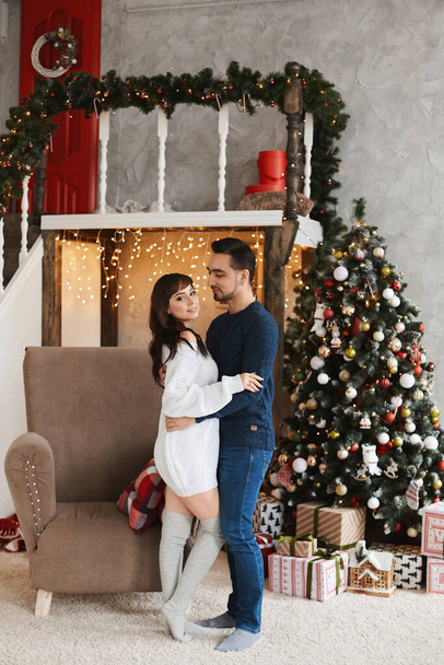Happy couple of young lovers hugging while posing near Christmas tree in the living room decorated for New year holidays - Fotoğraf, Görsel