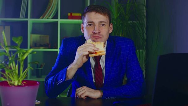 Young man eats sandwich, sitting in office. Businessman enjoying food at modern workplace during work. - Zdjęcie, obraz