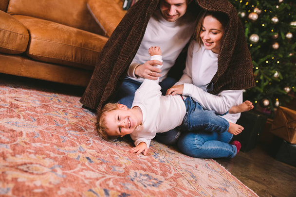 Thema Weihnachten und Neujahr in der Familie mit kleinem Kind. Dreiköpfige Familie sitzt am Vorabend von Weihnachten in eine Decke gehüllt auf dem Fußboden im Wohnzimmer neben dem Weihnachtsbaum. - Foto, Bild