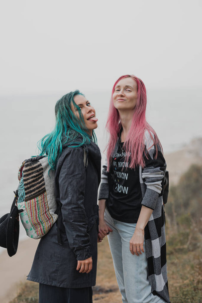 two women with pink and blue hair are holding hands and smiling while in the background there is a sandy beach and an ocean - Foto, imagen