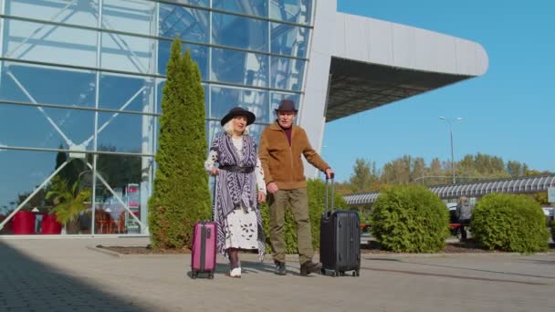Senior pensioner tourists grandmother grandfather walking from airport hall with luggage on wheels - Footage, Video