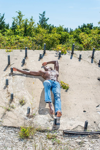 Bajo el sol, un hombre negro masculino, con pantalones vaqueros azules, descalzo y medio desnudo, está de pie en una antigua obra de construcción, con confianza mirando hacia adelante - Foto, imagen