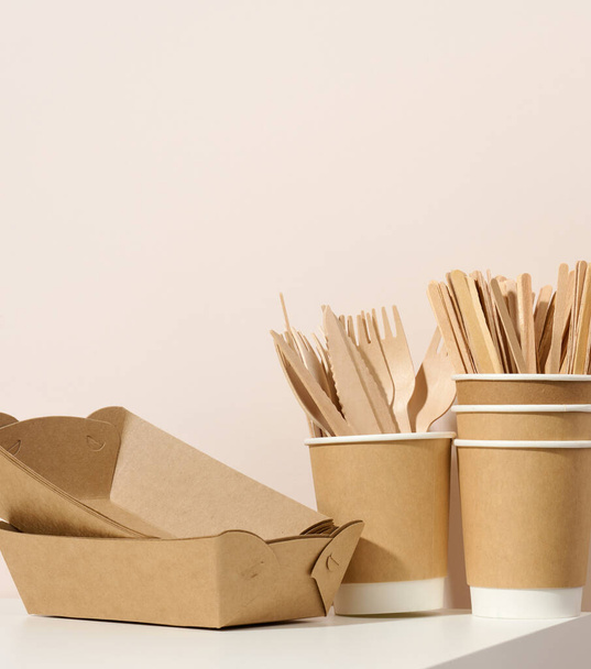 papel papelão pratos e copos castanhos, garfos de madeira e facas em uma mesa branca, fundo bege. Louça ecológica, sem resíduos - Foto, Imagem