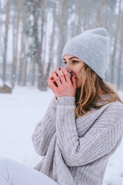Cute Caucasian girl in a white sweater and hat in a snowy forest Park drinking hot tea or chocolate from a thermos. Girl Wearing Warm Winter Clothes - Photo, Image