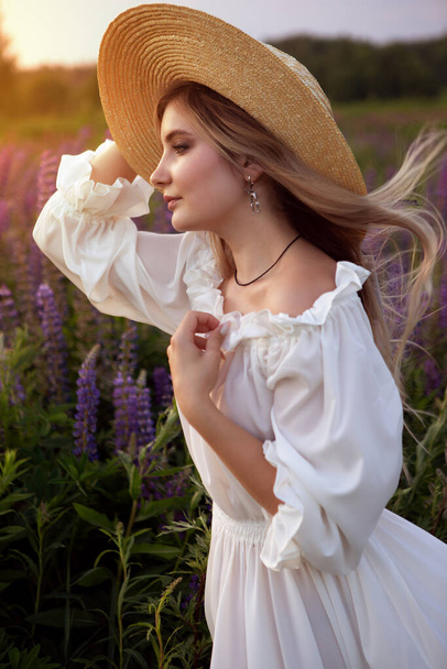 een modieuze foto van een mooi meisje met blond haar in elegante witte kleren en een strohoed, poserend op een zomerbloeiveld tijdens zonsondergang - Foto, afbeelding