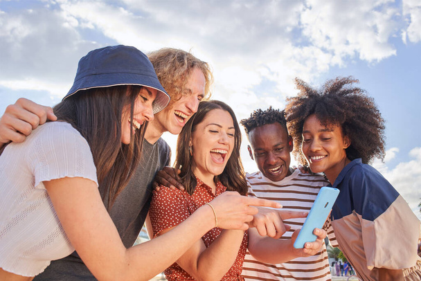 Eine Gruppe junger Menschen mit gemischter Rasse blickt auf ein Handy und zeigt auf etwas Lustiges auf dem Bildschirm. Konzept von Technologie, Verbindung, App, Online, Social Media. - Foto, Bild